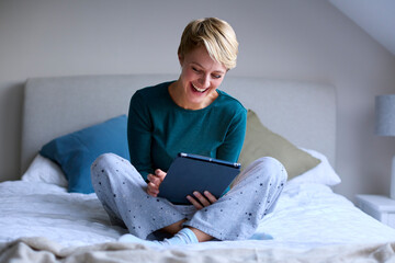 Young Smiling Woman Sitting On Bed Wearing Pyjamas At Home Looking At Digital Tablet