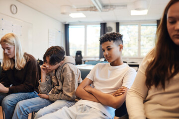 Thoughtful teenage boy sitting with arms crossed amidst friends in classroom during group therapy