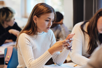Teenage girl with brown hair using smart phone sitting by female friend at desk in classroom