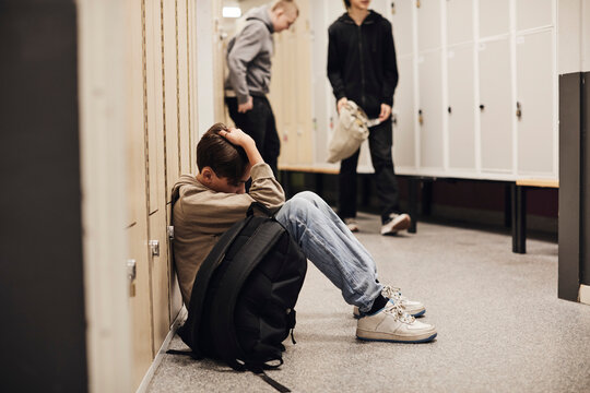 Full length of depressed boy sitting with backpack in school corridor