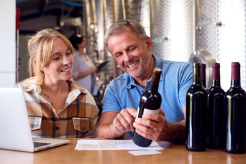 Man And Woman Checking Label On Bottle Inside Winery With Storage Tanks