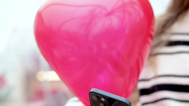 woman sitting in on table with paper hearts garland in one hand using smartphone with another,girl hugging red balloon heart shape.celebrate love valentine day coffee shop inside transparent balloon - Powered by Adobe