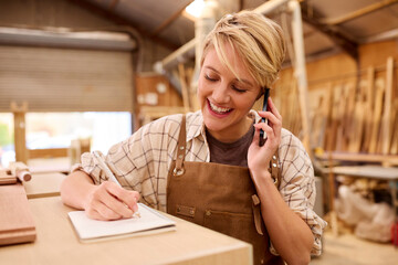 Female Apprentice Working As Carpenter In Furniture Workshop Making Phone Call