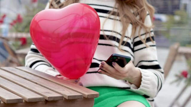 woman sitting in on table with paper hearts garland in one hand using smartphone with another,girl hugging red balloon heart shape.celebrate love valentine day coffee shop inside transparent balloon