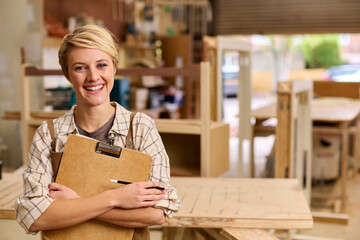 Portrait Of Female Apprentice With Clipboard Working As Carpenter In Furniture Workshop 
