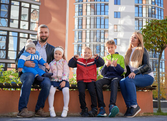 Fototapeta premium Happy family - father, mother and children having fun together on playground. Parents and kids sitting on bench, looking to camera. Modern residential buildings on background.