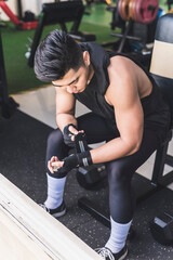 A buff asian guy adjusts his gloves while sitting down on the bench. Working out at the gym.