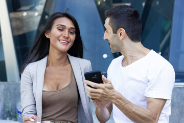 Caucasian man showing viral video on phone to Asian woman colleague in urban city. Multiracial teamwork funny business talk, startup project ideas, modern technology concepts