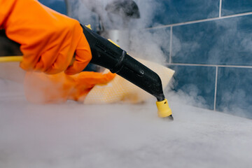 A woman's hand in an orange rubber glove washes the tile in the kitchen using a steam generator. Professional cleaning service. The concept of cleaning the bathroom or kitchen, cleanliness.