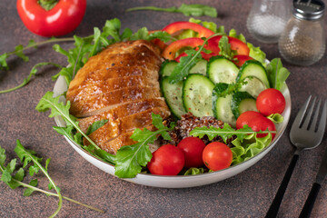 Salad with baked chicken breast, quinoa, cherry tomatoes, cucumbers, sweet pepper, lettuce leaves and arugula on brown table