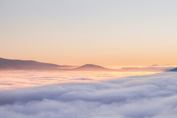 View of sea of clouds colored in the soft orange-pink hue of the morning sun. Peaks of the...