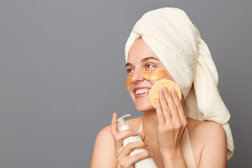 Closeup portrait of joyful attractive Caucasian young woman wearing towel holding sponge and lotion in hands, looking away with satisfied facial expression, enjoying morning routine.