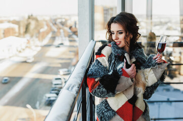 a sexy girl dressed in a long fur coat poses on the balcony against the background of the winter city; holding a glass of red wine