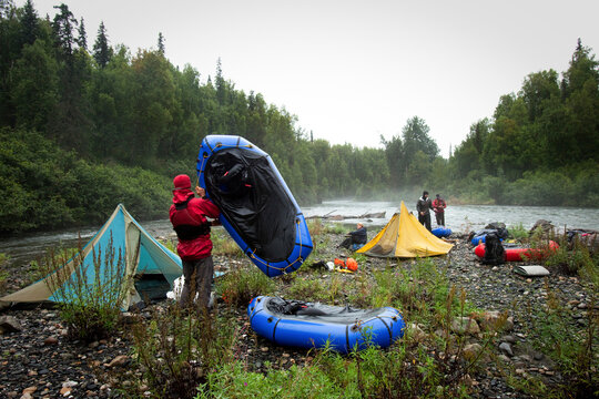 Four friends packing up camp by a river with tents and packrafts in the rain.