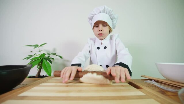 5 years old caucasian boy dressed like a chef rolls out pastry with a rolling pin cooking cookies or a pie or a pizza. A boy prepares dough for baking in the oven