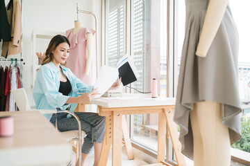 Portrait of a confident Asian tailor woman working on clothes in tailoring atelier. A young attractive female fashion designer smile and look at the camera with a dress-on mannequin in the background.