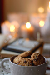 Cup of tea, books, tablet, bowl of cookies, various spices, pine cones and lit candles. Hygge at home. Selective focus.