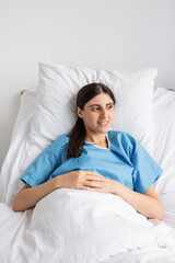 Positive woman in patient gown lying on bed in clinic.