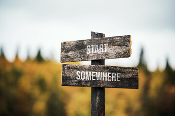 vintage and rustic wooden signpost with the weathered text quote start somewhere, outdoors in nature. blurred out forest fall colors in the background.