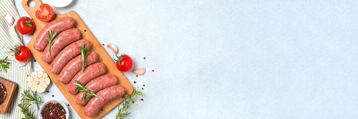 Long banner with raw sausages or bratwurst with ingredients ready to cook on wooden cutting board over white stone table background with copy space. Traditional pork sausages. Overhead view