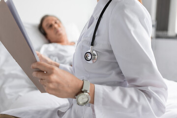 Doctor in white coat holding paper folder near blurred patient in hospital ward.