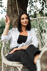 Beautiful happy girl in fashionable casual clothes with a white shirt sits and rides on a knitted vintage swing in the park