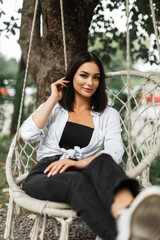 Style beautiful happy girl business lady in fashionable casual clothes with a white shirt sits and rests on a knitted swing
