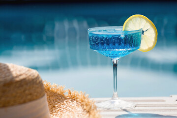 A glass of delicious blue cocktail Blue Lagoon and yellow lemons on the background of the pool. Alcoholic cocktail juicy fruit blue with curacao liqueur, ice cubes and a slice of lemon. - obrazy, fototapety, plakaty