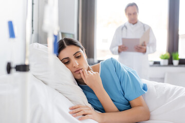 Obraz premium brunette woman in patient gown lying on bed near blurred doctor in clinic.