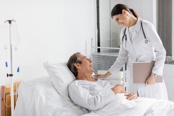 Side view of smiling doctor with paper folder calming elderly patient in hospital ward.