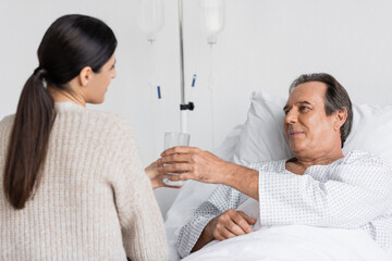 Fototapeta premium Blurred daughter giving glass of water to senior dad on hospital bed.