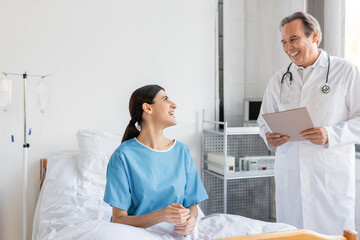 Fototapeta premium Smiling senior doctor holding paper folder near patient in hospital ward.