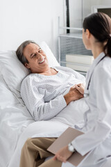 Blurred doctor holding hand of smiling senior patient in clinic.