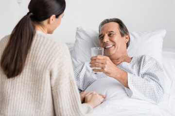 Obraz na płótnie Canvas Smiling senior patient holding glass of water near blurred daughter in clinic.