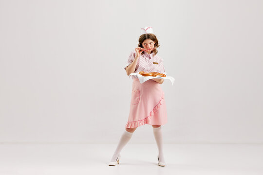 Beautiful young girl in image of retro cater waiter wearing 70s, 80s fashion style uniform isolated over light studio background. Charm, ad, sales, fashion, emotions