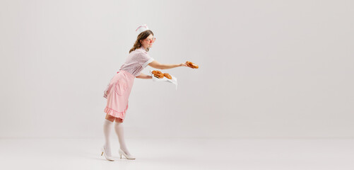 Portrait of young female waitress, sensual woman in retro american fashion style of 70s, 80s treats with croissants over grey background.