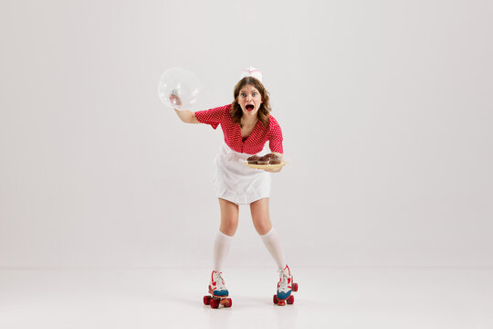 Emotional excited young girl, stylish retro waitress in american fashion style of 70s, 80s uniform rollerblade over gray background. Art, creativity, emotions, service concept