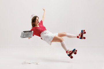 Emotional excited young girl, stylish retro waitress in american fashion style of 70s, 80s uniform rollerblade over gray background. Falling - obrazy, fototapety, plakaty