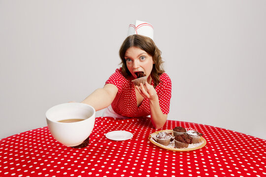 Stylish young girl in retro american fashion style of 70s, 80s uniform sitting at table and tasting cupcakes with coffee over grey background. Pop art Photography