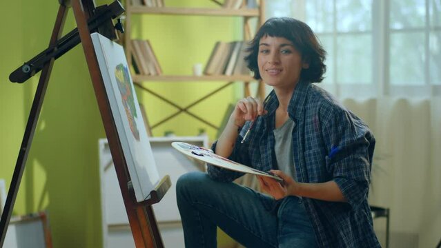 A beautiful woman wearing a blue flannel t shirt is painting something amazing and modern on her canvas in a big and brightly lit art atelier