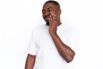 Irritated young man suffering from toothache. Male African American model with closed eyes, short haircut and beard in white T-shirt touching his cheek having toothache. Pain, disease concept