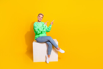 Full body photo of pretty cheerful lady sit podium indicate fingers empty space isolated on yellow color background