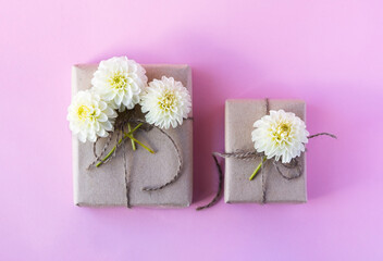 Obraz na płótnie Canvas Two gift boxes in gray craft paper decorated with white dahlia flowers on a pink background for the holiday.