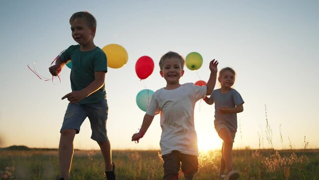Child birthday. Girls and boy play in park at sunset in grass. Family run with balloons. Happy cheerful children outdoors in field. Summer in park smile children with balloons. Happy childhood concept