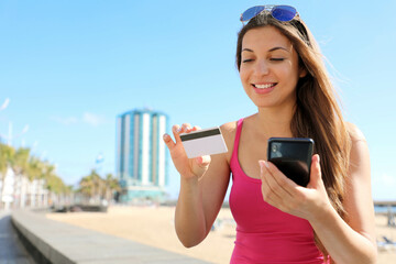 Excited young woman buying online with mobile phone reading credit card number outdoor on summer