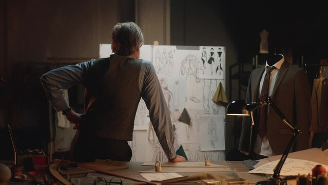 Male mature tailor works in luxury designer atelier or tailoring studio. He looks at board with sketches of future clothes and thinks. Mannequin with tailored suit. Fashion and hand craft concept.