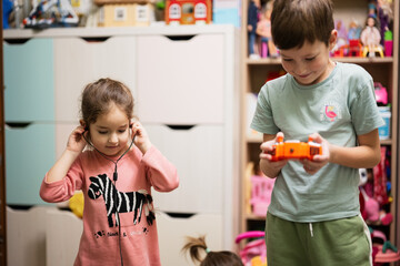 Brother hold remote control and sister listening to music through wired headphones in children's room.