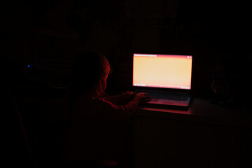 Baby girl tap on laptop while watch on colored screen in light off room.