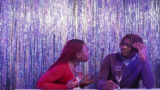 Bad party. Quarreling couple. Problem relationship. Angry black woman screaming on displeased careless man sitting on shimmering cascade curtain background.