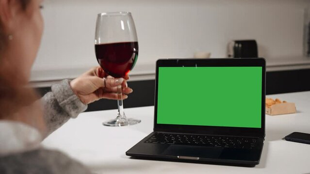 woman drinking red wine looking on display laptop with green screen on the table in kitchen evening at home